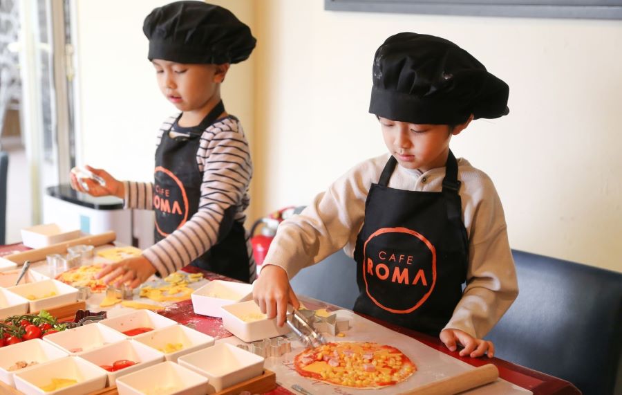 Two young boys in Cafe Roma's black chef's hats and aprons make pizza as part of a workshop organised by the restaurant. They have the pizza bases in front of them and are adding toppings to them.
