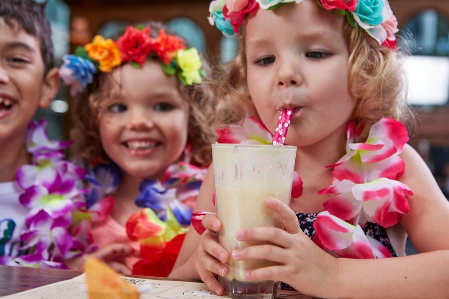 A child wearing a garland and flower crown drinks a milkshake at Frites. To her left, two other smiling children watch her.