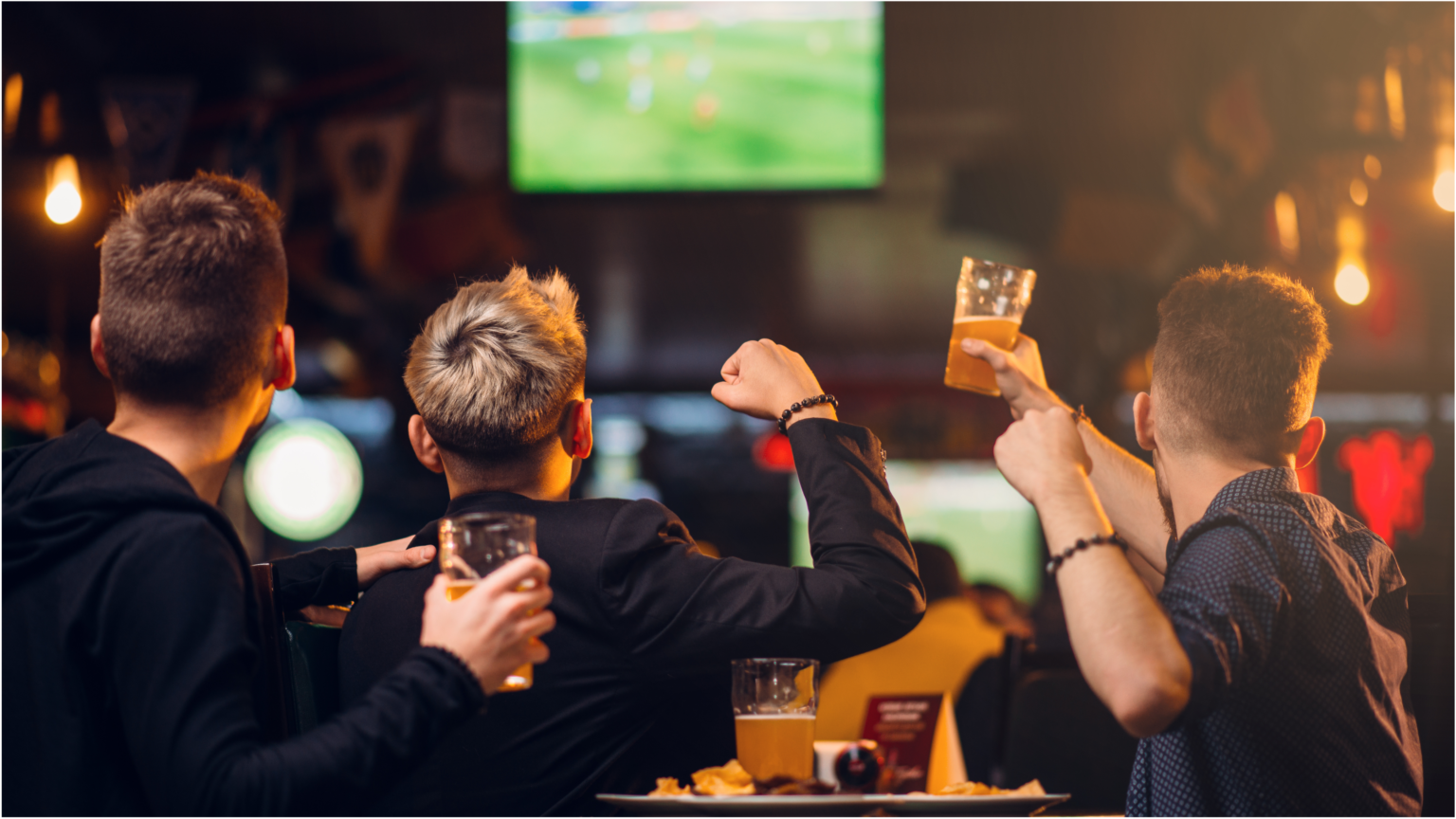 Football fans cheer as they watch a game at a pub and raise their glasses.