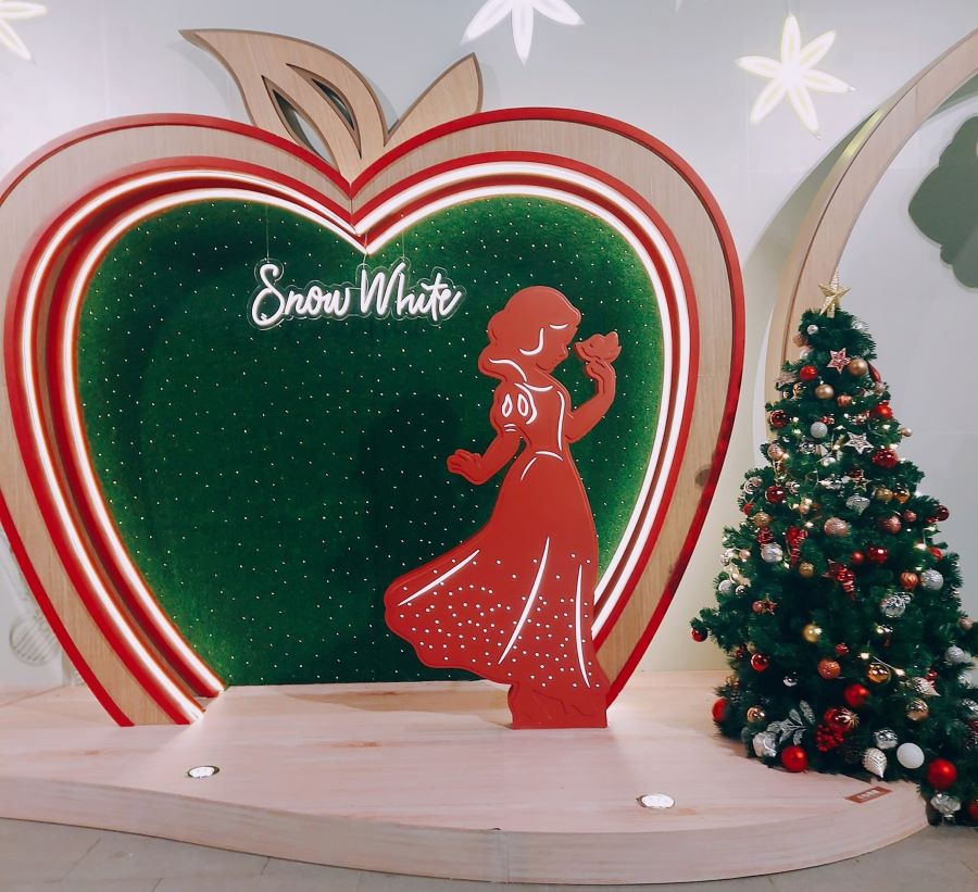 The Snow White display at Tsuen Wan Plaza’s Green Fantasy Christmas. There is a red silhouette of Snow White in front of an apple-shaped photo booth. There is a Christmas tree on the right side of the picture.