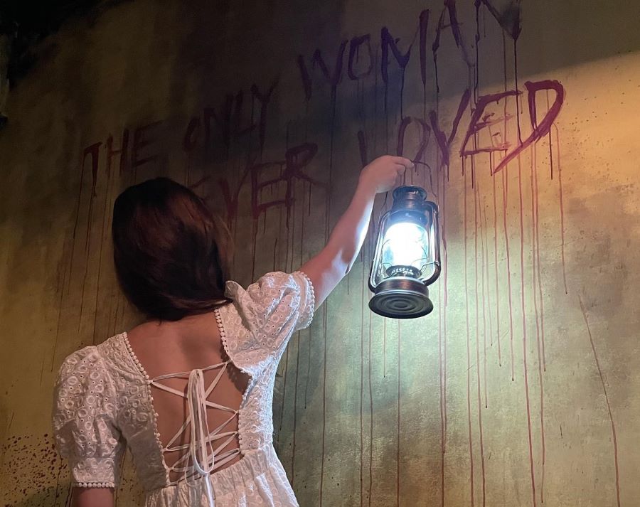 A woman in a white dress holds up a lantern to see the writing on a blood-stained wall.