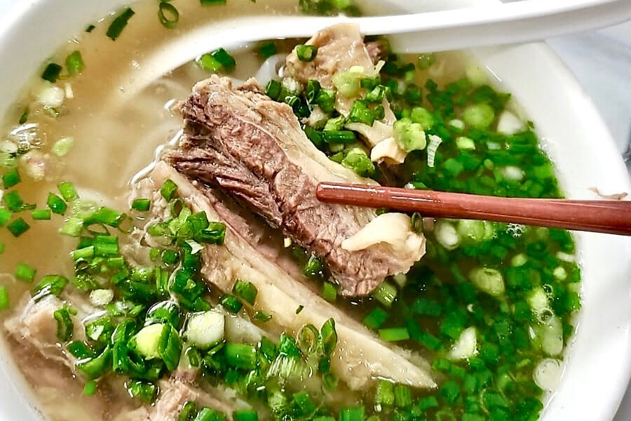 a pair of chopstick holding a piece of tender beef brisket in clear broth and chinese chives