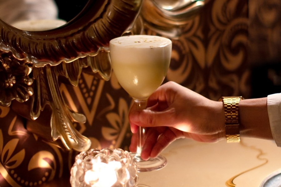 In a corner of The Wise King bar, a person holding a cocktail with opulent wallpaper and golden framed mirror in the background