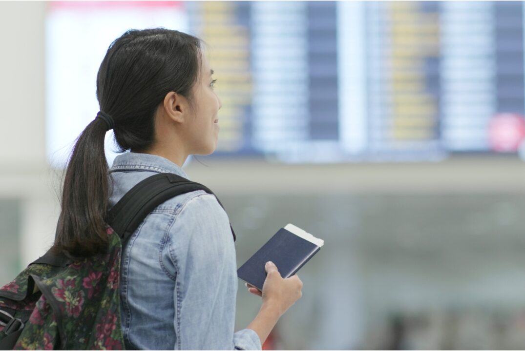 A woman at the departures hall of the Hong Kong International Airport. She is holding her passport, wearing a blue collared shirt, and a blackpack.