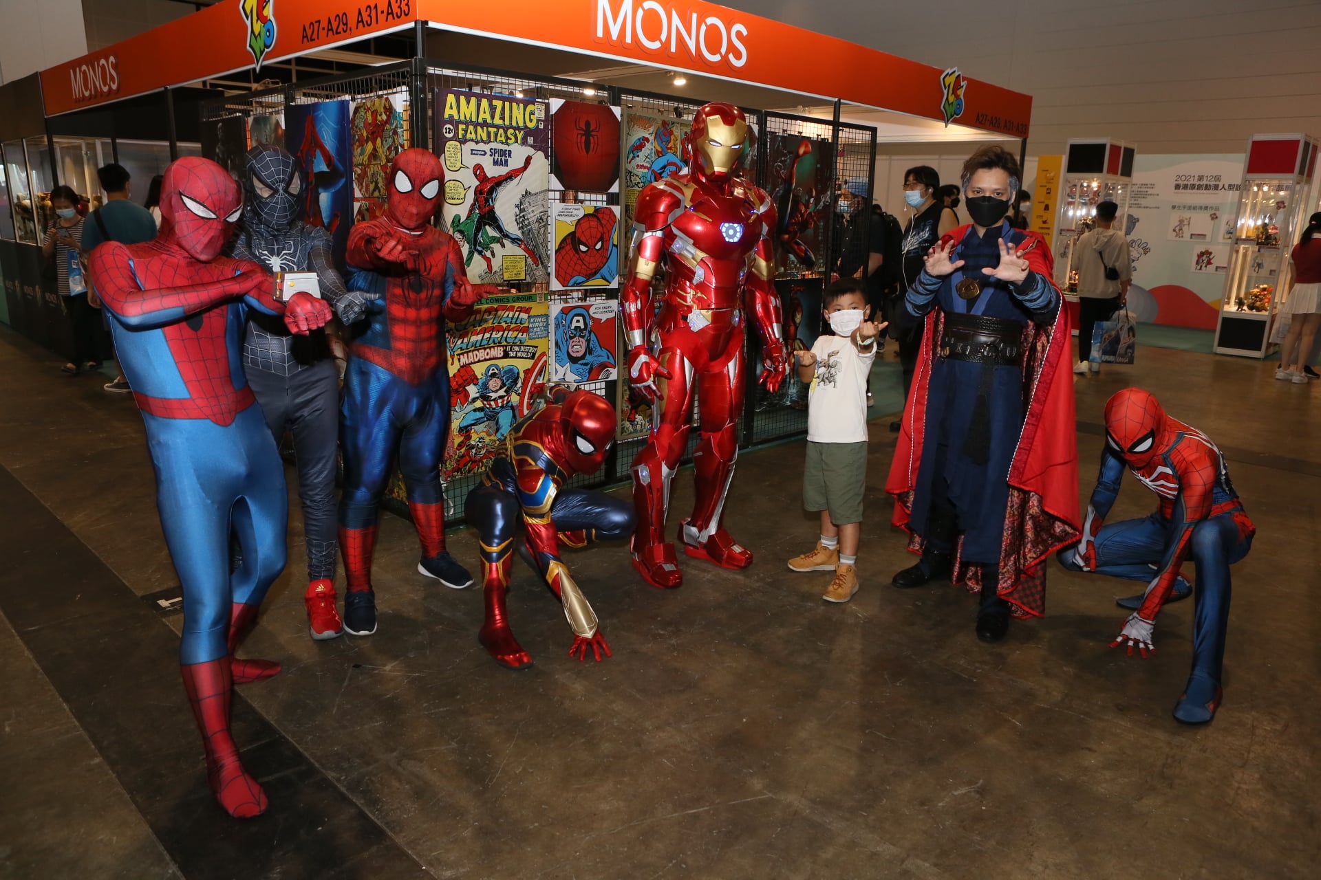 A group of 8 people pose in front of stalls at Ani-Con & Games Hong Kong. A child in a white t-shirt and olive green shorts (third from left) poses with cosplayers dressed as various versions of Spider-Man, Iron Man and Doctor Strange.