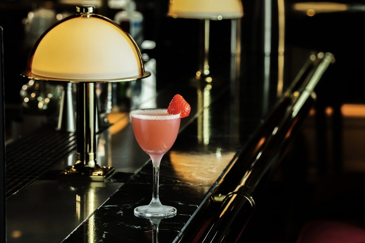 A cocktail glass containing a pink drink and strawberry sits on a dark counter. The counter is lined with low lamps woth dome-shaped shades.