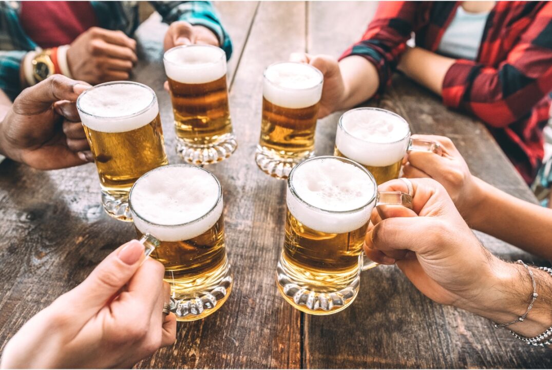 A group of six friends, faces unseen, put their mugs together before drinking craft beer. They are sitting at a wooden table.