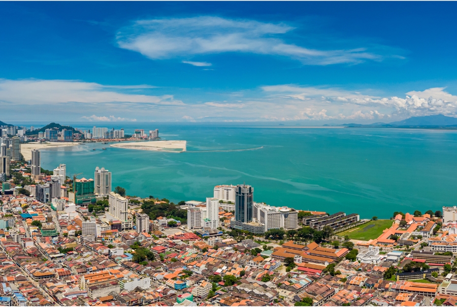 An aerial view of Georgetown in Penang, Malaysia. The cityscape is a combination of high-rises and low-rise tiled buildings. In the background, there are sandy beach shores.