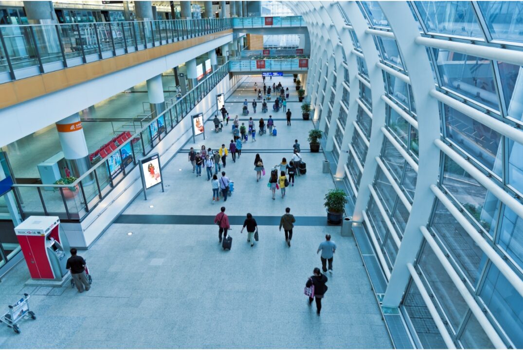 An overhead view of an entry hall in the Hong Kong International Airport. Several people walk up and down the hall carrying luggage or bags.