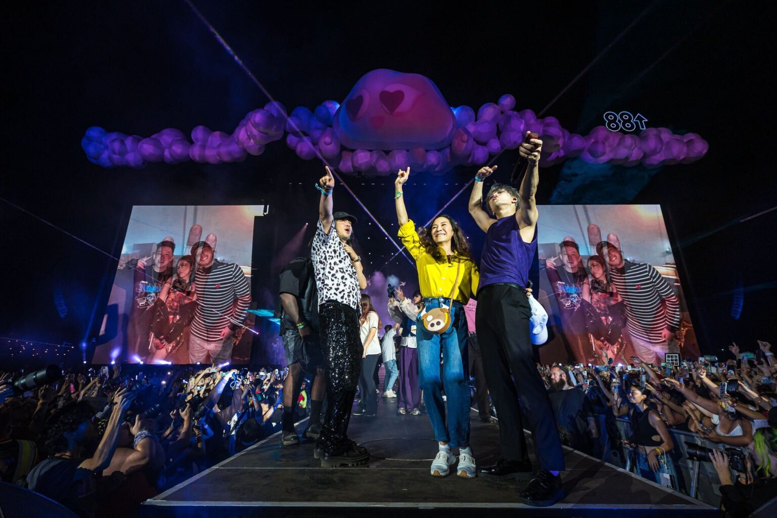 Rich Bran, Michelle Yeoh, and Jackson Wang on the Head In The Clouds stage in Los Angeles. The trio is flanked by screens and crowds, and the festival's trademark clouds are suspended above them.
