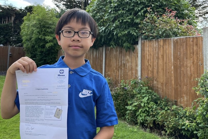 Adrian Li, an 11-year-old boy, wears a blkue shirt and glasses, holding up a paper. The paper is a document from the Mensa Society congratulating him on his high score in the test.