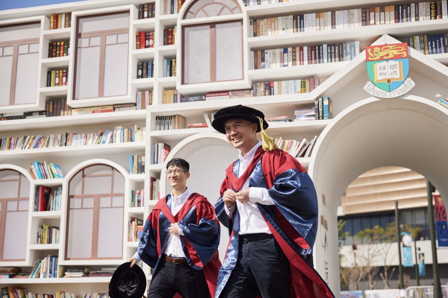 Two students wearing graduation gowns in front of university bookshelves lined with books.