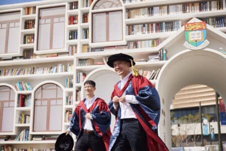 Hong Kong Goes Up 3 Spots In International Rankings To Become World’s 12th Best Student City Two students wearing graduation gowns in front of university bookshelves lined with books.