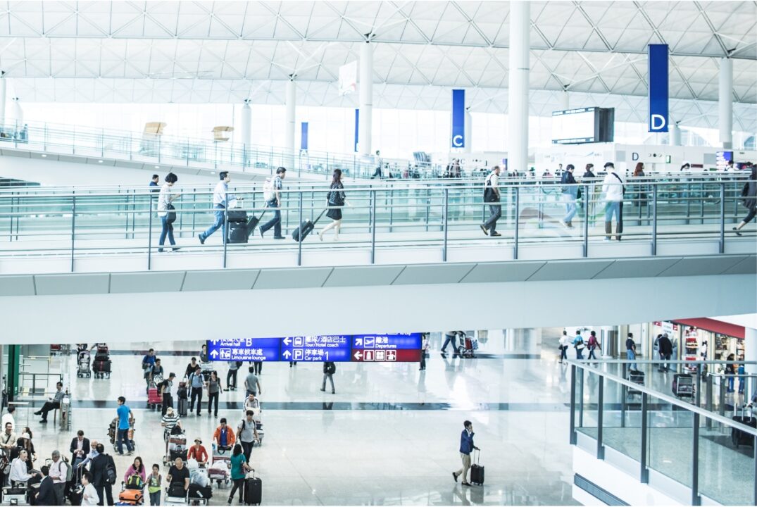 An image of the Hong Kong International Airport. There is a passage on which passengers walk to the departures hall. The arrivals hall, which has people walking up and down it, is on the level below.