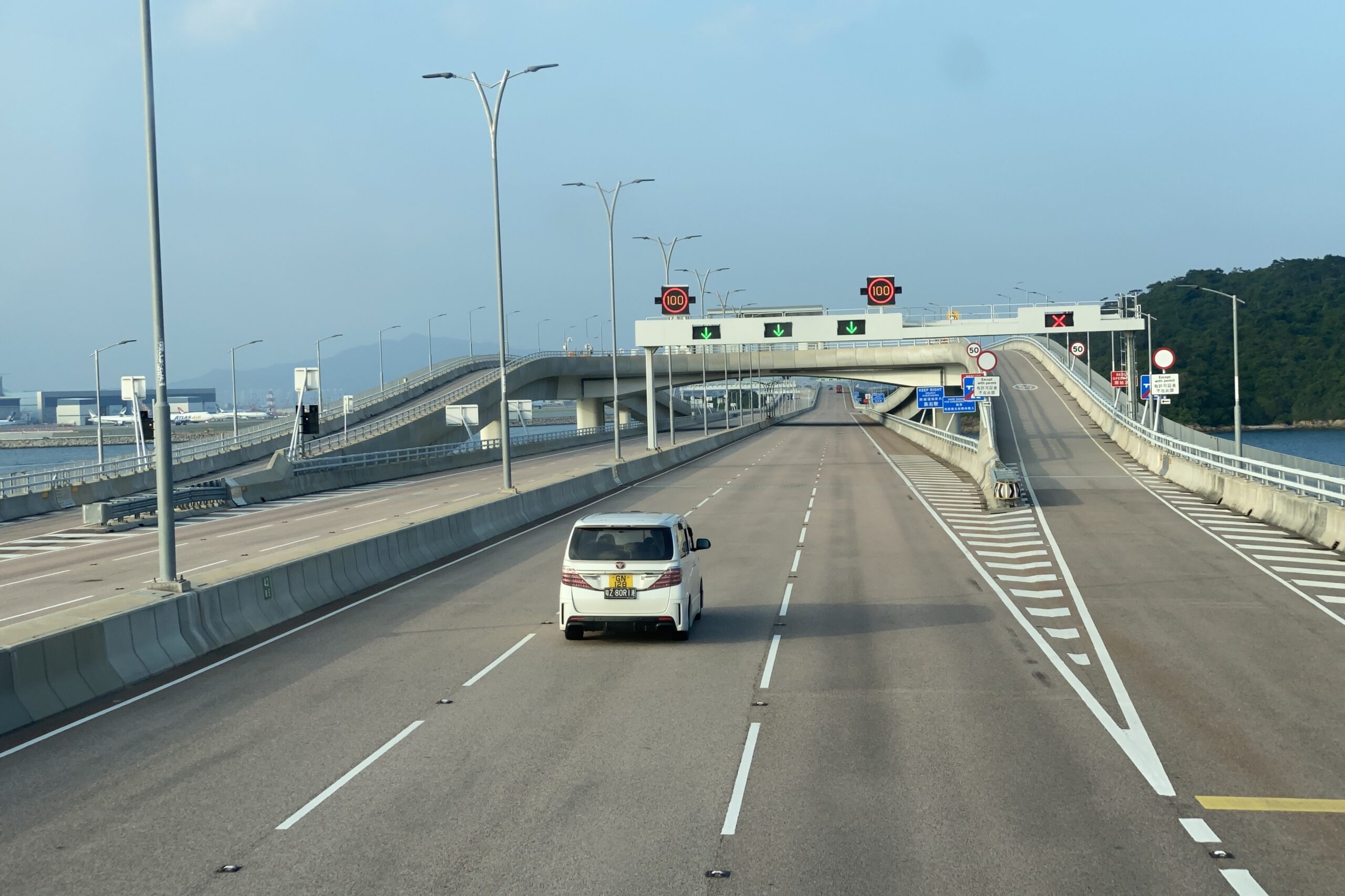 A white car drives down the Hong Kong section of the six-lane Hong Kong-Zhuhai-Macao Bridge. There are two indicators of the 100km/hr speed limit, as well as green arrow signs showing where cars should drive.