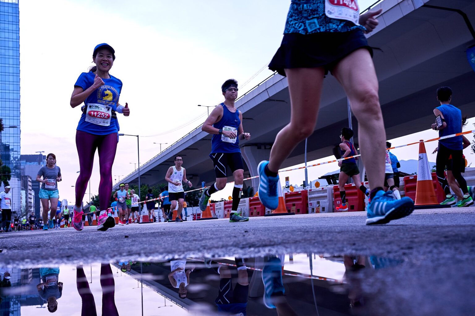 A low-angle shot of runners at the Hong Kong Streetathon. Some of the runners' reflections can be seen in a puddle as they run with an elevated expresswayin the background.