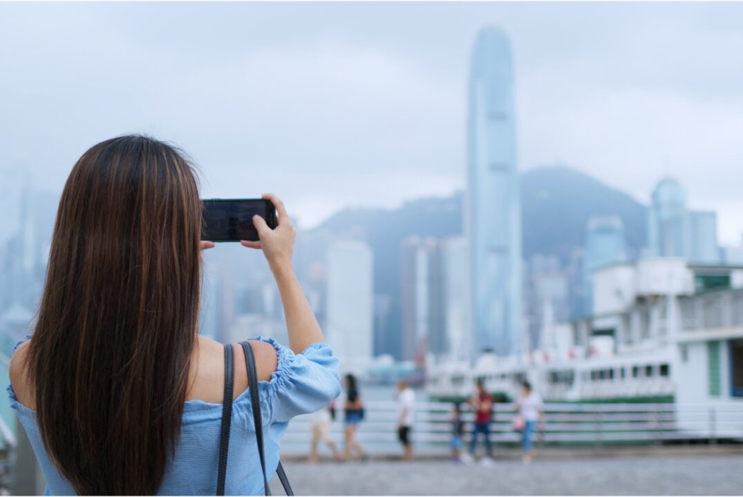 A woman in a blue off-shoulder top with long dark brown hair, takes a picture of IFC in Hong Kong using her mobile phone. The woman is on the Kowloon side of Victoria Harbour.
