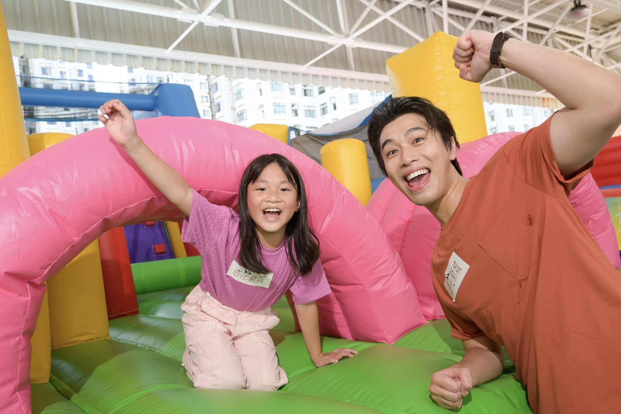 A girl and man pose in front of a colourful inflatable obstacle course. They are cheering with their hands.