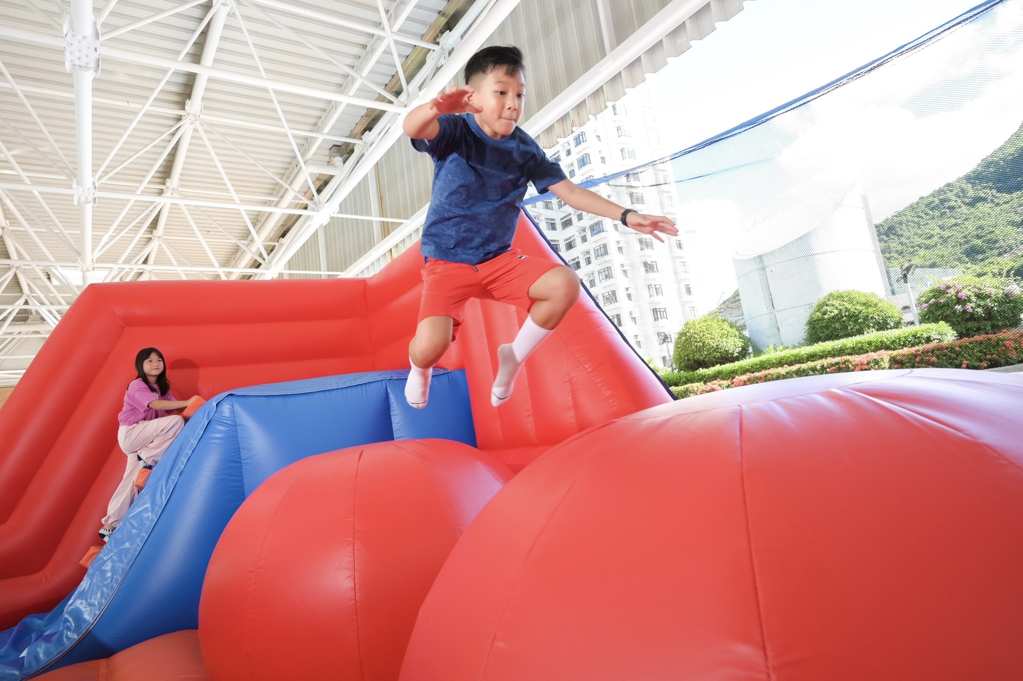 Two children play on an inflatable platform with a ladder and two giant balls. A girl is in the background, climbing the ladder. A boy is jumping from one ball to the one in front of him. 