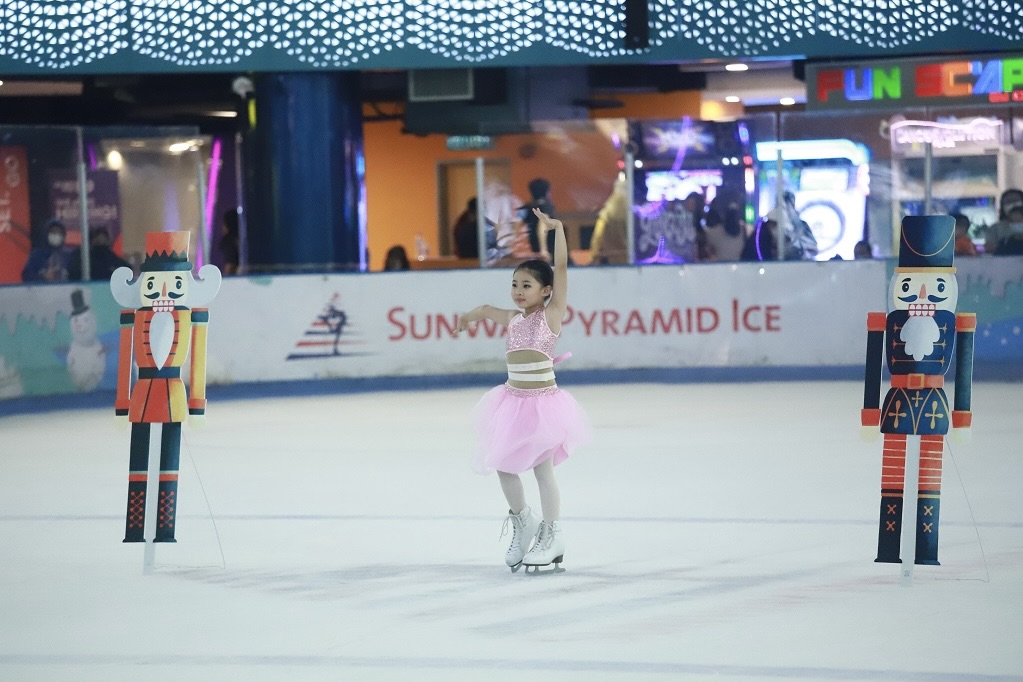 A child in a figure skating outfit stands on an ice skating rink. She is flanked by cardboard cutout soldiers.