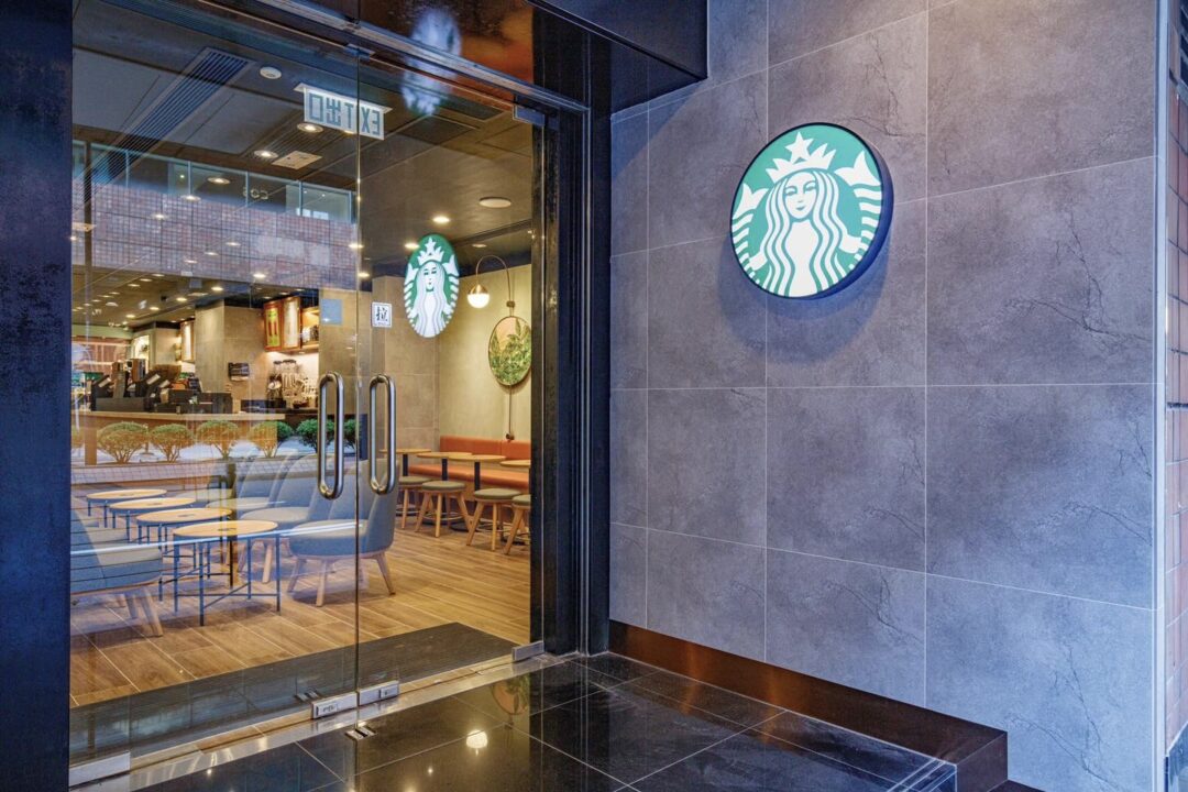The exteriors of a Starbucks outlet in Hong Kong. The Starbucks logo adorns a granite walls, and a glass door on the left leads into the coffee shop. Through the glass door, one can see chairs, tables, and a counter.