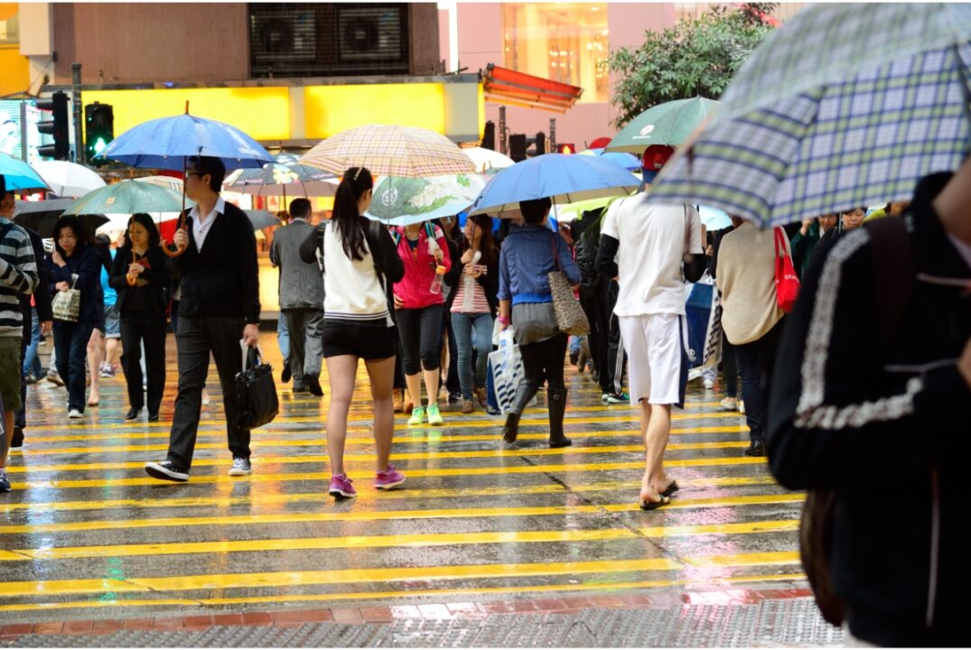 Pedestrians holding umbrellas cross a busy street in Hong Kong. The street is wet with rain.