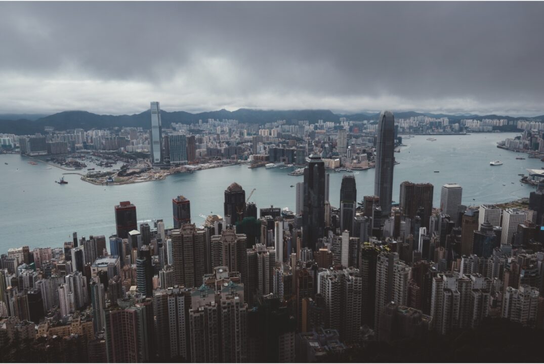 An overhead view of Victoria Harbour in Hong Kong. In the foreground is Hong Kong Island with Kowloon in the background. The sky is overcast and grey.