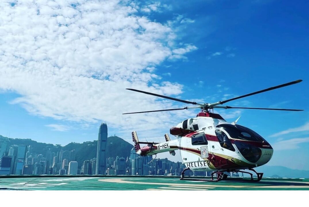 A helicopter on a helipad on the Kowloon side of Victoria Harbour. The skyscrapers on Hong Kong Island are visible in the background. There sky is a clear blue with a scattering of white clouds.