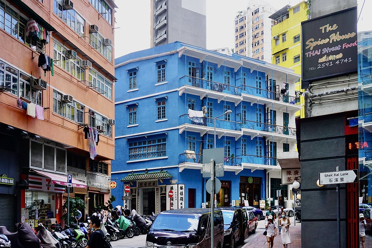 The Blue House on Stone Nullah Lane flanked by the Orange House on its left and the Yellow House on the right. The street in front of the houses has parked scooters and cars, and pedestrians walk on the pavements. 