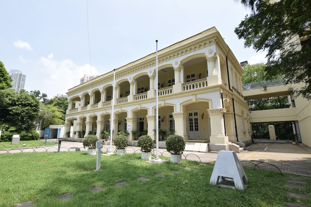 The original Hong Kong Observatory building is a two-storeyed brick plaster building with arched windows and long verandas. It has a lawn in front of the entrance.