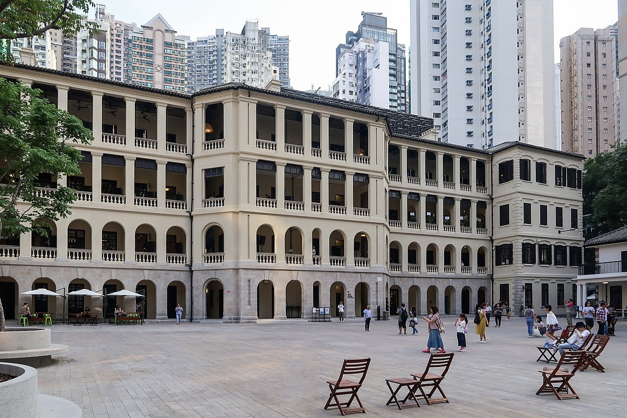The exterior of Tai Kwun, which is distinctive for its long corridors and verandas. The courtyard in front of the facade has chairs that passers-by can sit on. People are walking through the courtyard, and some stand and take pictures.