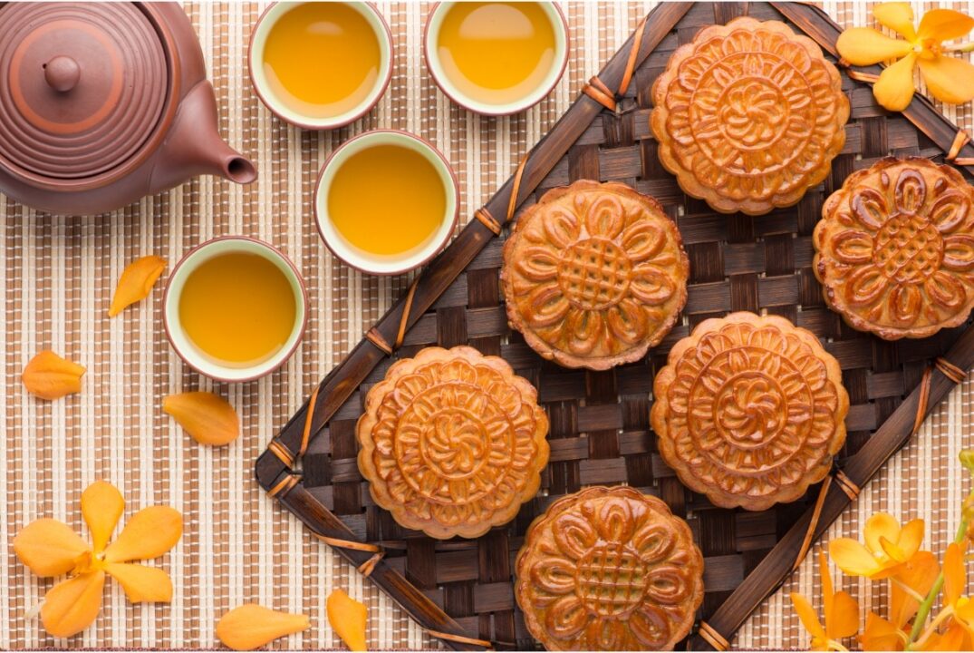 An overhead view of a tray of 6 mooncakes sitting in a 2 x 3 configuration. There are 4 cups of tea to the left of the tray, as well as a teapot. They all sit on a striped tablecloth that is strewn with yellow flower petals.