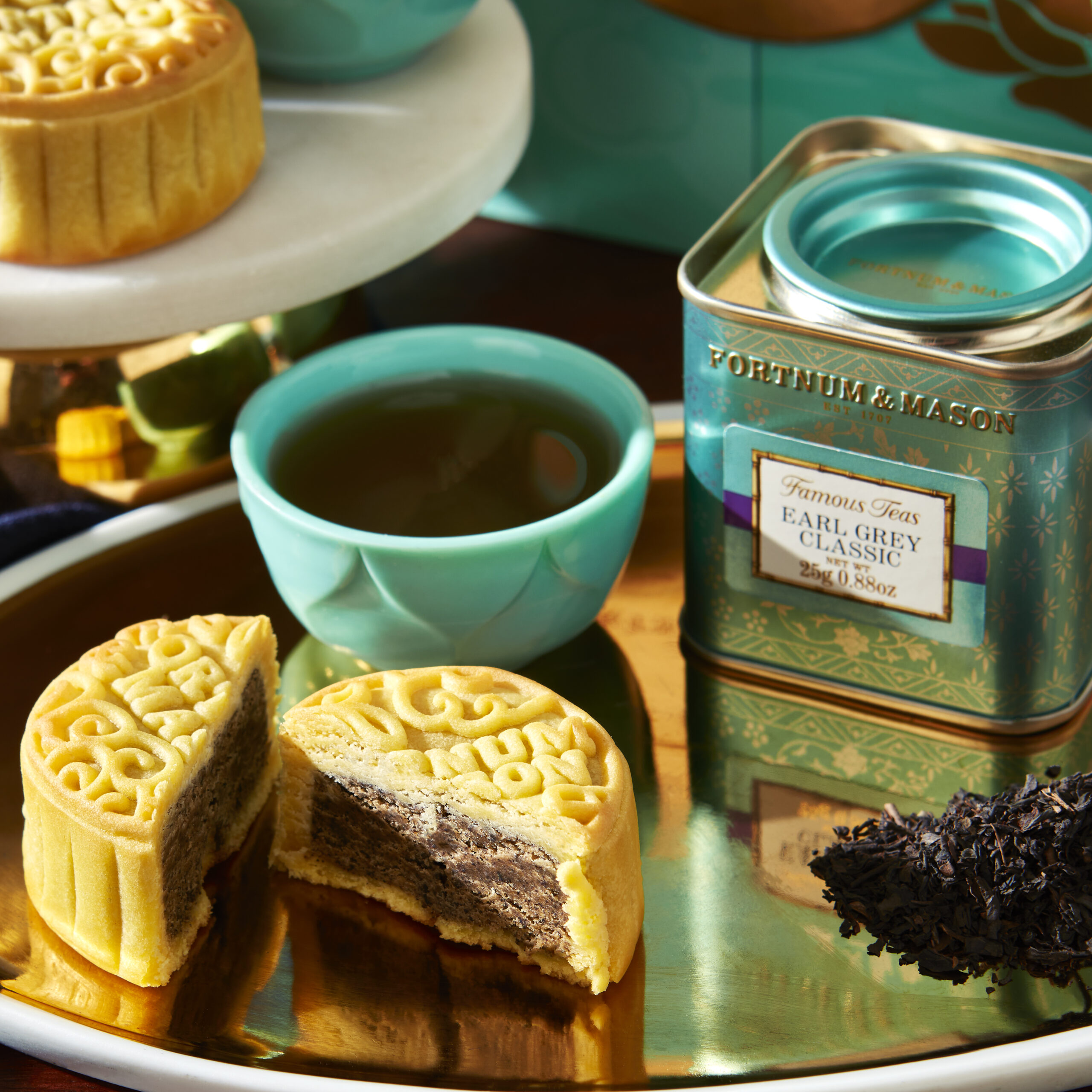 A light golden brown mooncake from Fortnum & Mason cut into half to reveal its filling sits on a tray, along with a teal cup of tea, a matching teal tin box, and tea leaves. In the background, there is a whole mooncake sitting on a stand, which is partially visible with a giftbox in the background. 