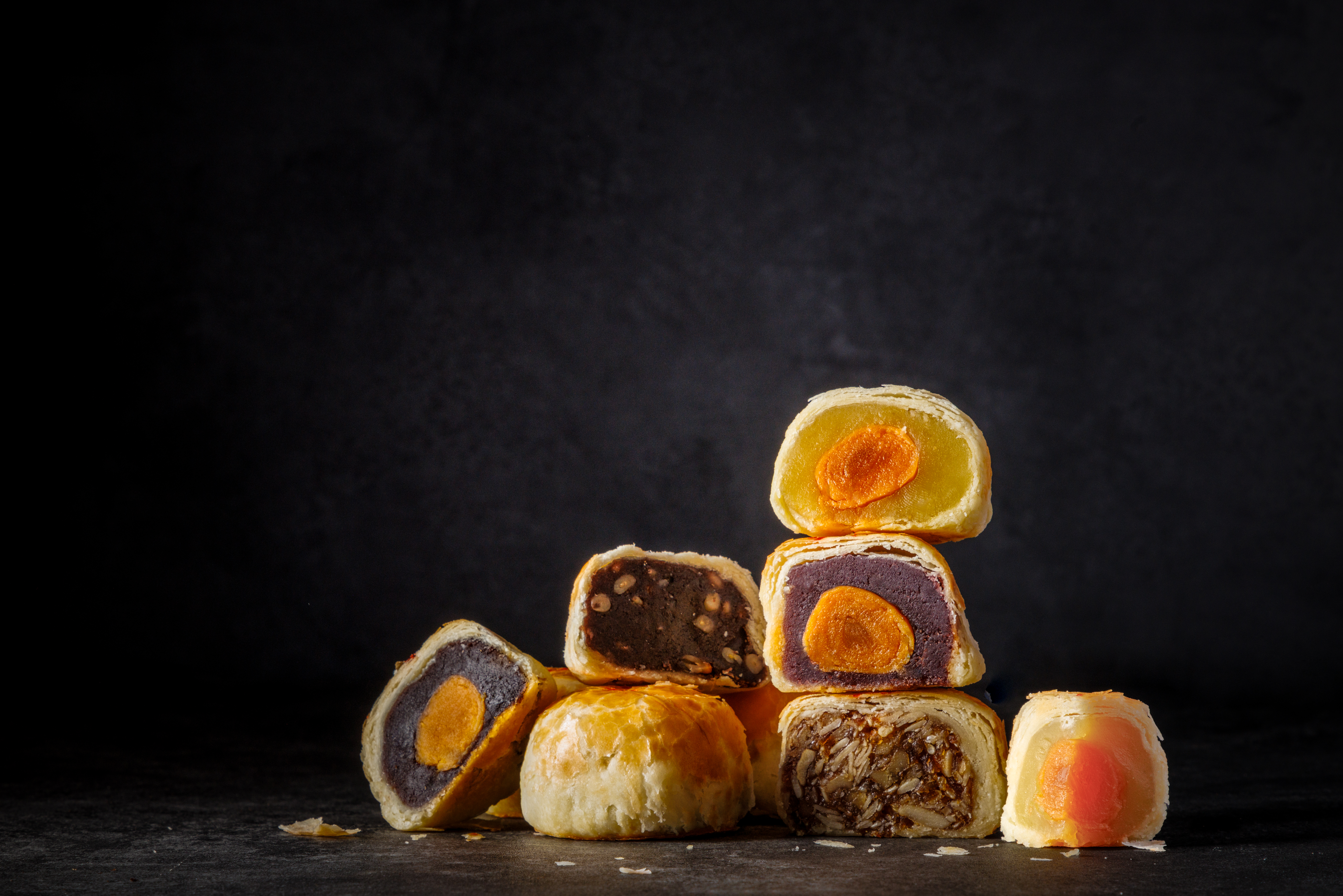 Six different mooncakes cut in half to reveal their fillings sit in a pile on a dark table with a dark background. There is one half of a mooncake at the bottom that shows only the pastry half.