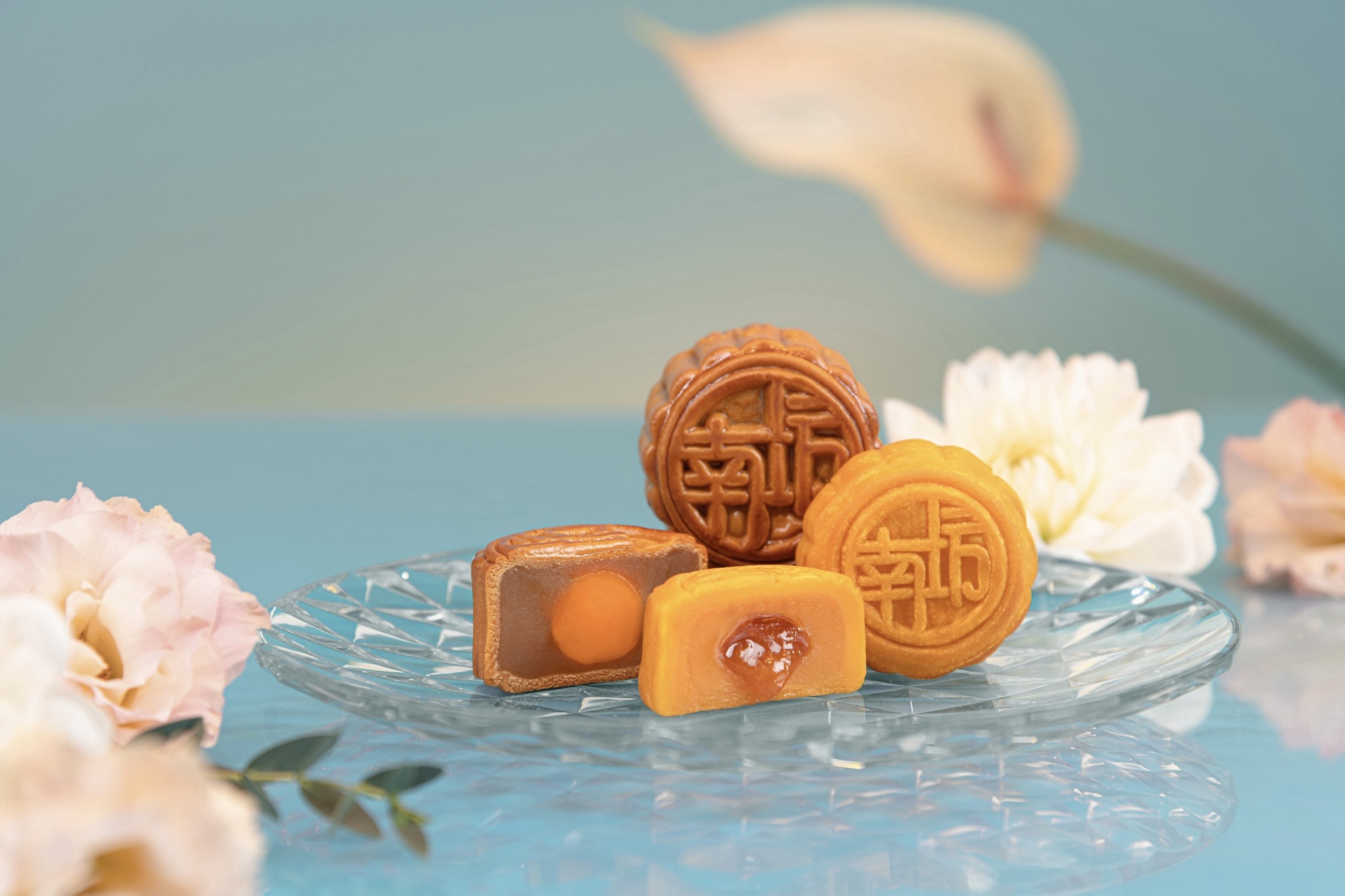 Two whole mini mooncakes sit on a glass plate, along with two mini mooncakes cut in half to reveal their fillings. The darker brown mooncakes at the back are the lotus seed paste with egg yolk, and the lighter brown ones in the front are the lava egg custard ones. The mooncakes are surrounded by flowers and placed on a blue table top.
