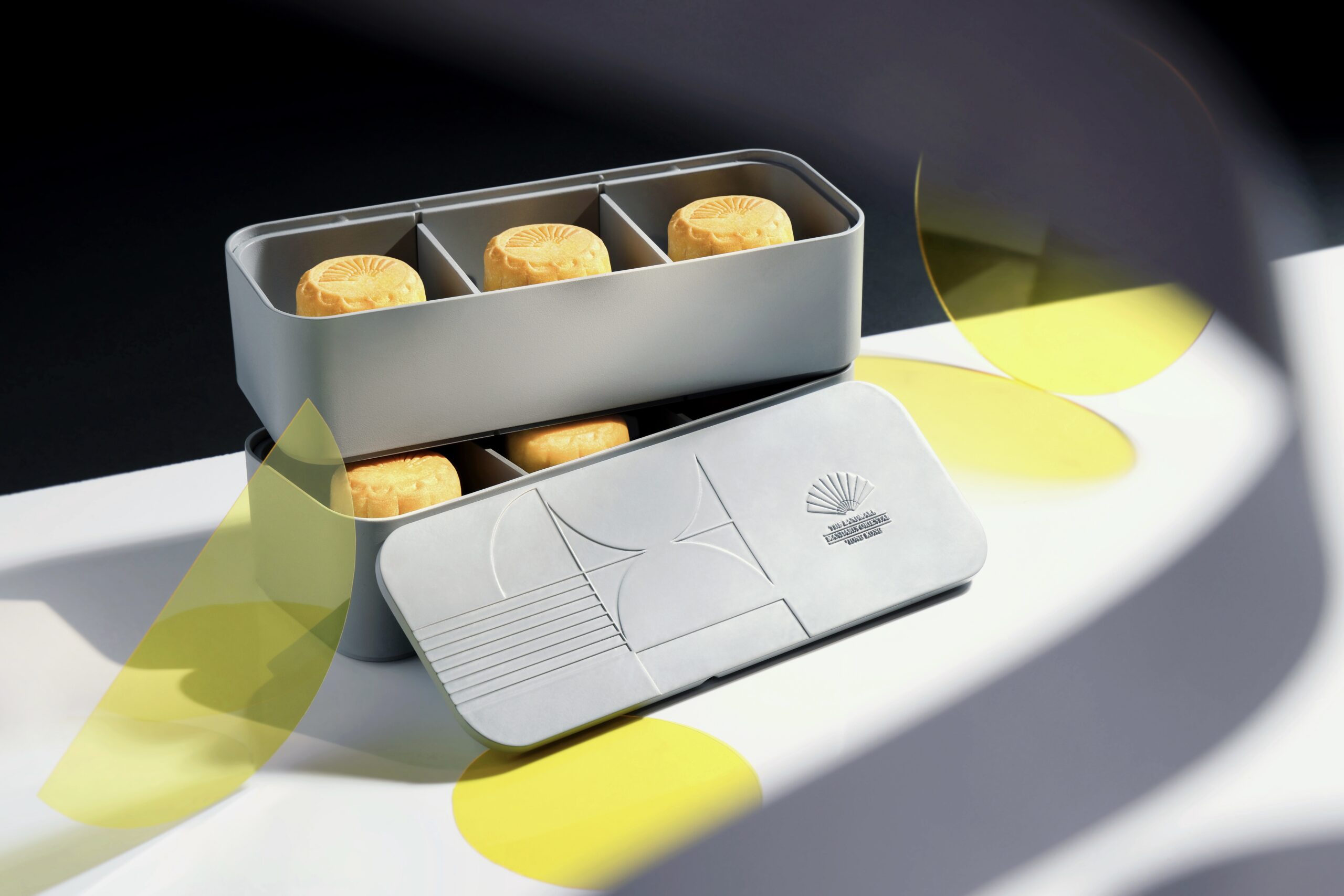 A light grey double-decker partitioned bento box with mooncakes sits on a table. The top level of the box is completely uncovered to reveal three mooncakes within. It sits upon the lower level of the bento box, which contains partially obscured mooncakes. The lid of the box is propped against the bottom level to show the logo of the Landmark Mandarin Oriental. There are neon yellow wrappers strewn around the box, which is placed on a white table top.