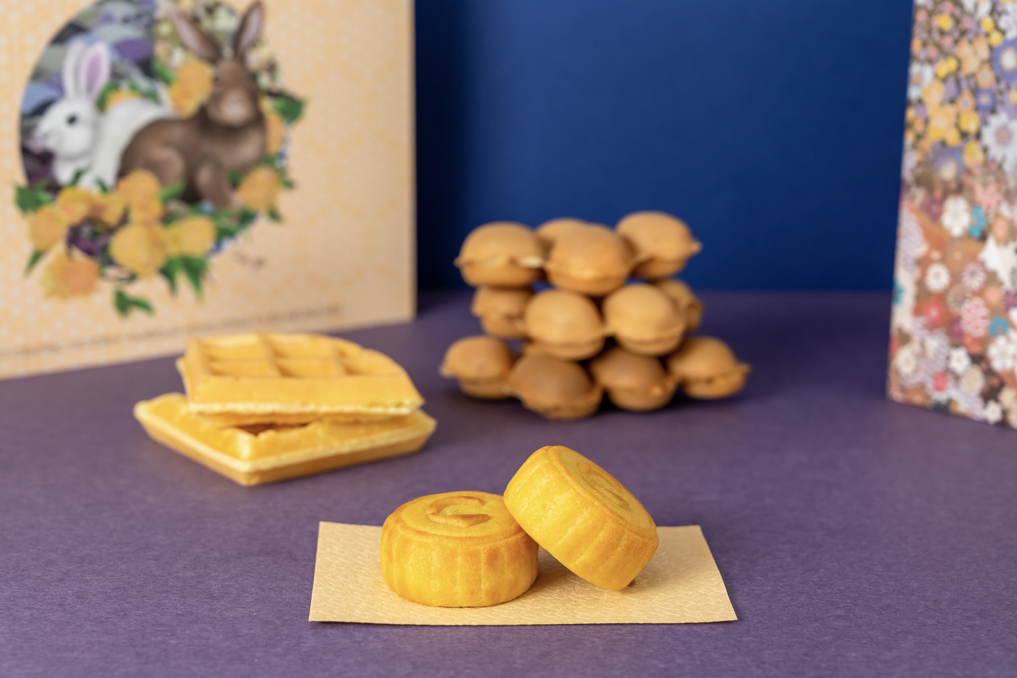 Two whole mooncakes sit placed on top of the other. In the background, there is a traditional waffle and a bubble waffle, as well as a box with rabbits.