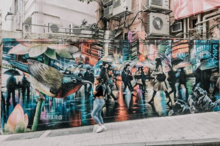 A woman walks past a mural depicting an oversized bird watching people walk down a street as they hold umbrellas.