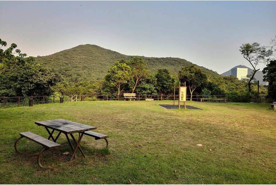 A bench at Aberdeen Country Park in Hong Kong. There is exercise equipment and a fence in the background, as well as verdant hills.