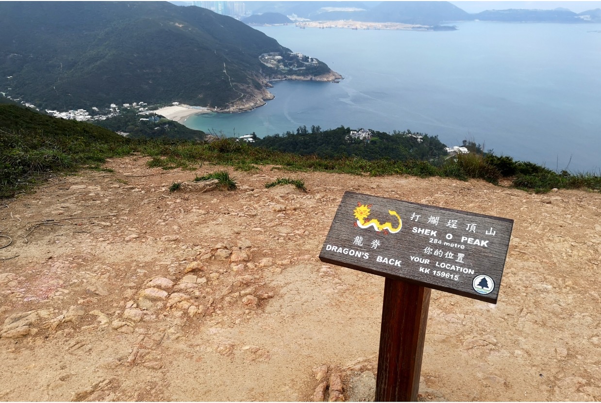 A sign showing the height of Shek O Peak on Dragon's Back Trail. The peak overlooks the sea and a beachy area with an urban settlement.