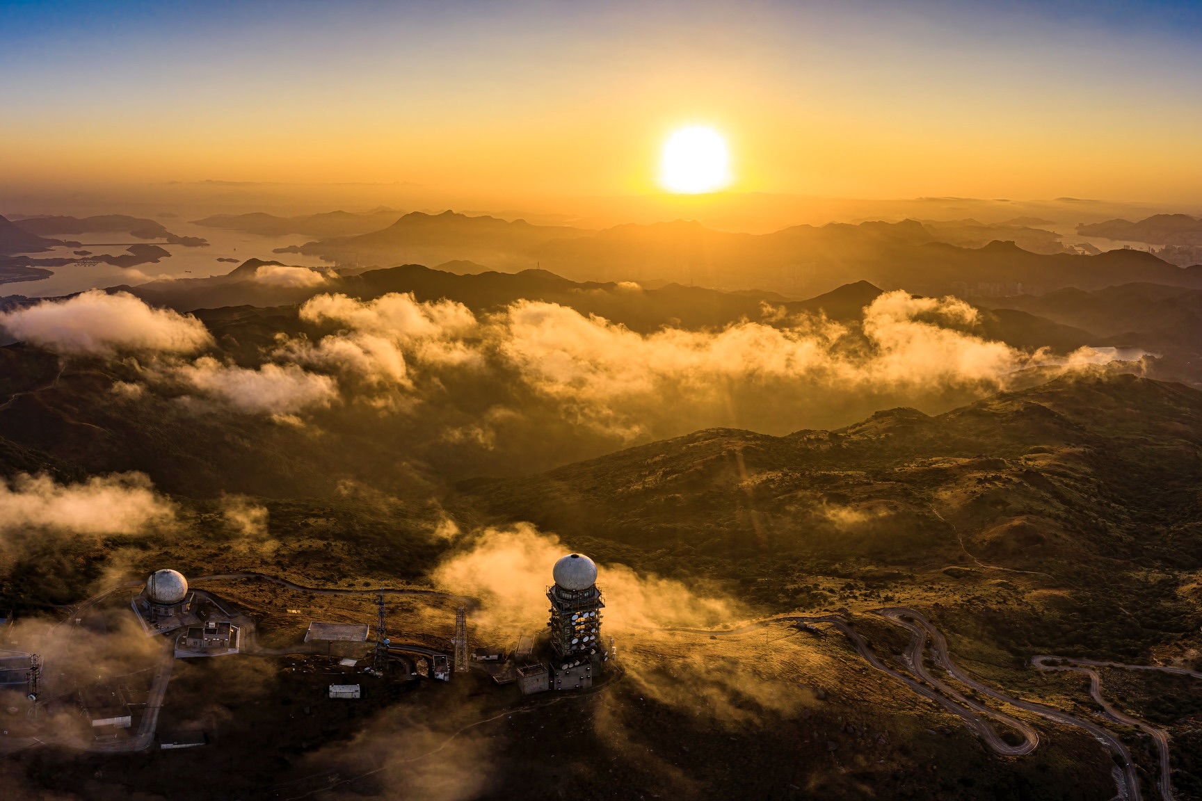 An overhead view of the Hong Kong Observatory's weather radar station in Tai Mo Shan Country Park at sunrise. The rolling hills are blocked by low-lying clouds.