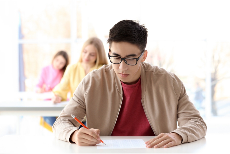 A male student does a multiple-choice exam. He is wearing a light brown jacket over a red shirt, and glasses. He sits at a white table and holds an orange pencil. There are two female students behind him, also taking the exam.
