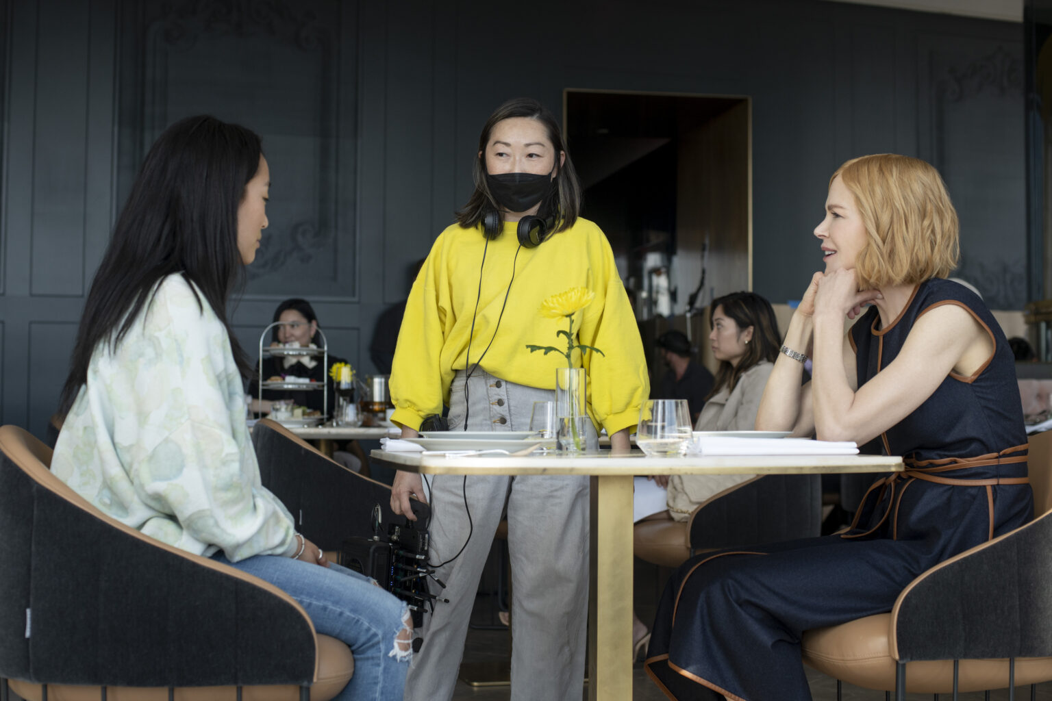 Ji-young Yoo, Lulu Wang and Nicole Kidman on the set of the web series Expats. You and Kidman sit at a table in a restaurant, while Wang stands and wears a face mask.