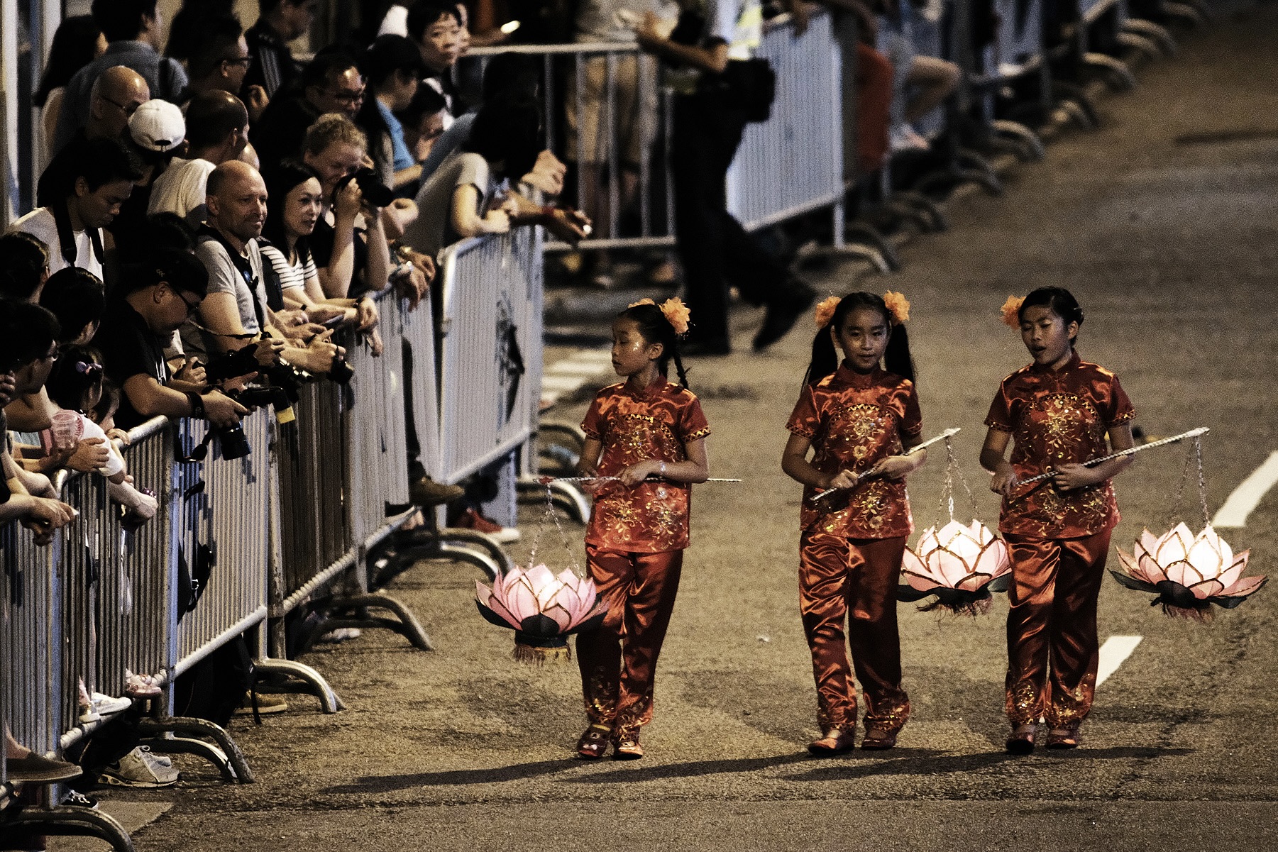 Three children wearing traditional Chinese outfits hold lotus-shaped lanterns suspended from rods and walk ahead of the fire dragon dance procession. A crowd of people from behind a barricade watch them.