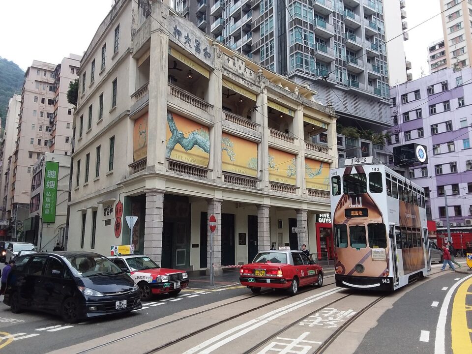 A view of Woo Cheong Pawn Shop. There is a tram and taxi in front of the building, as well as cars emerging on to the street in front of the building from a side road.