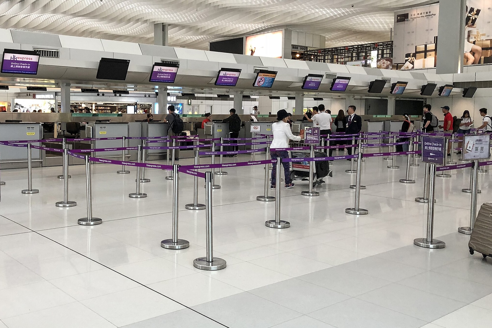 The HK Express check-in counters at Hong Kong International Airport. There are queue managers placed in front of the counters and passengers walking through them and standing in line waiting to check in,