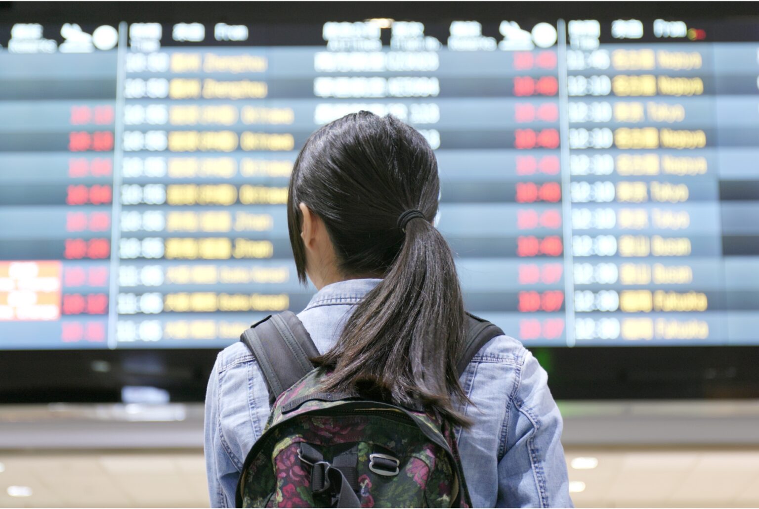 A woman looks at a departure board at the Hong Kong International Airport. Her back faces the camera as she looks at the board. She has her long hair tied back in a low ponytail that falls down her back. She wears a blue collared shirt and a backpack.