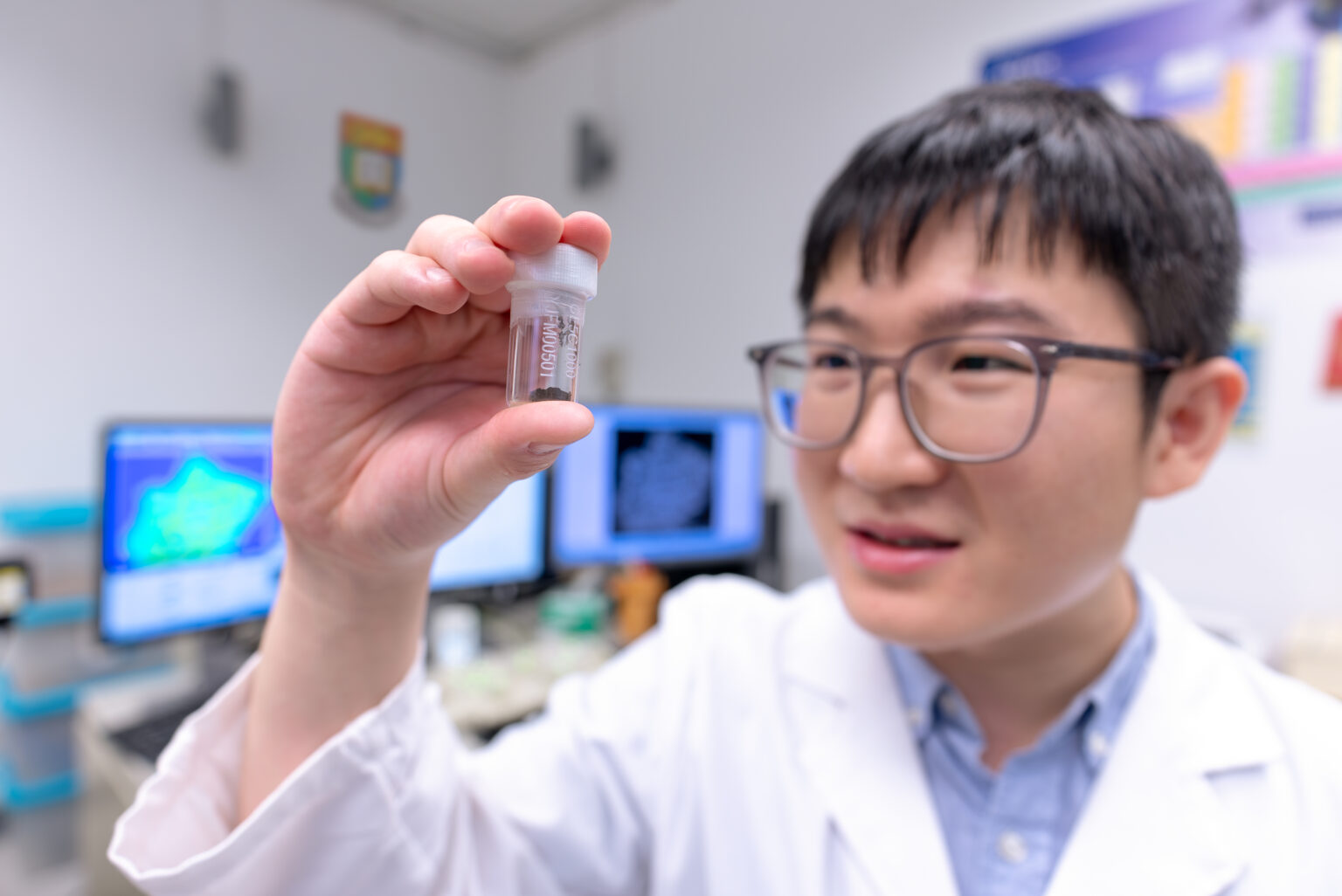 Dr Yuqi Qian from the University of Hong Kong holds up a container with lunar soil samples. The doctor is wearing a white lab coat with a blue shirt underneath. He is in a research lab with computers in the background.