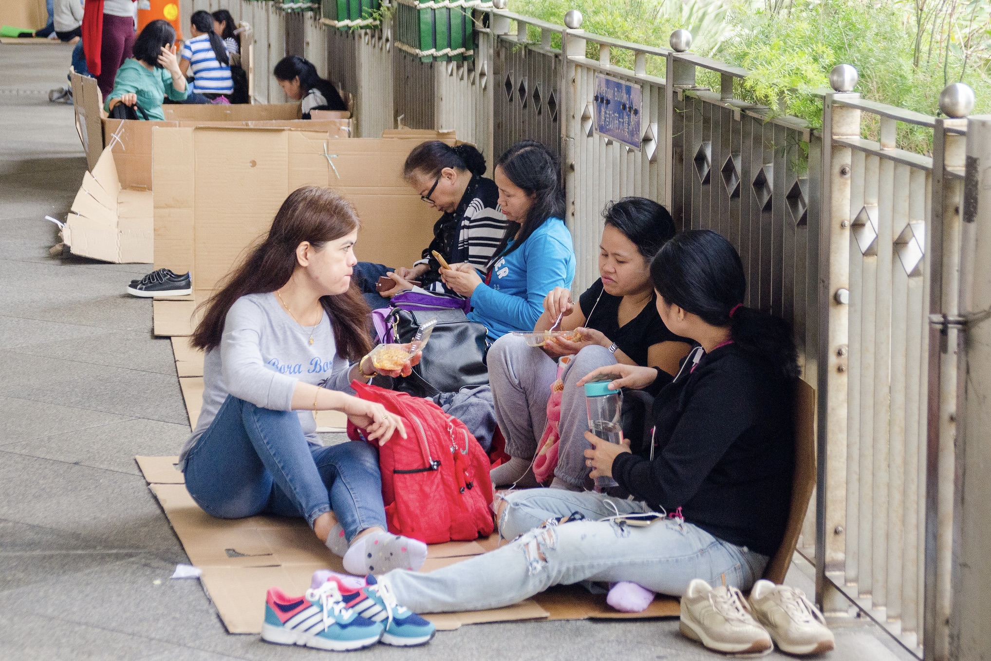 A group of foreign domestic helpers sit on flattened cardboard boxes at a covered pedestrian walkway in Hong Kong. They are relaxed and not wearing their shoes. Some of them are eating, while others scroll through their mobile phones.