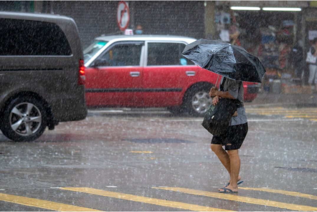 A woman walks in heavy rain with an umbrella. There are cars, most notably, a red Hong Kong taxi, in the background.